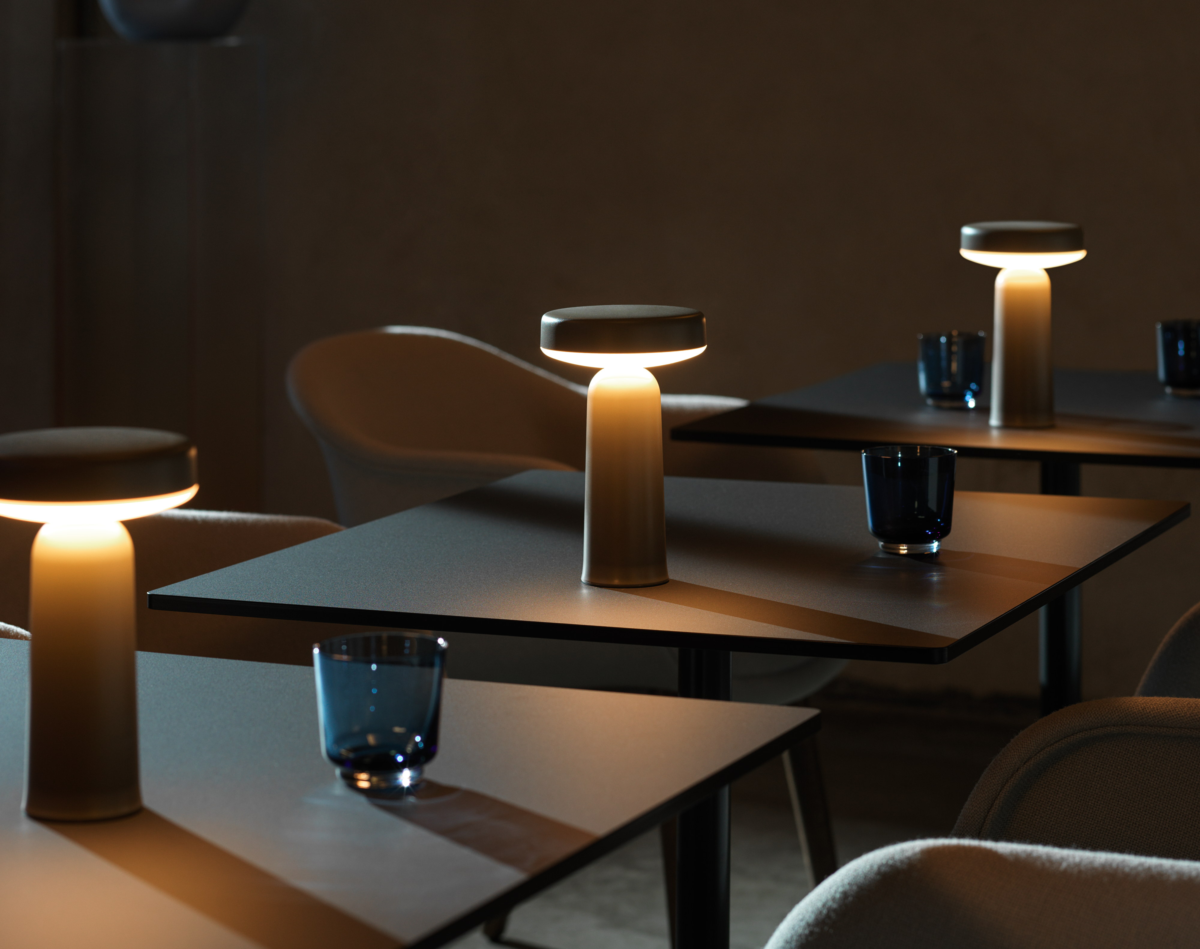 A dimly lit café setting with multiple Muuto Ease portable lamps casting a warm glow on dark tables, accompanied by blue drinking glasses and soft chairs.
