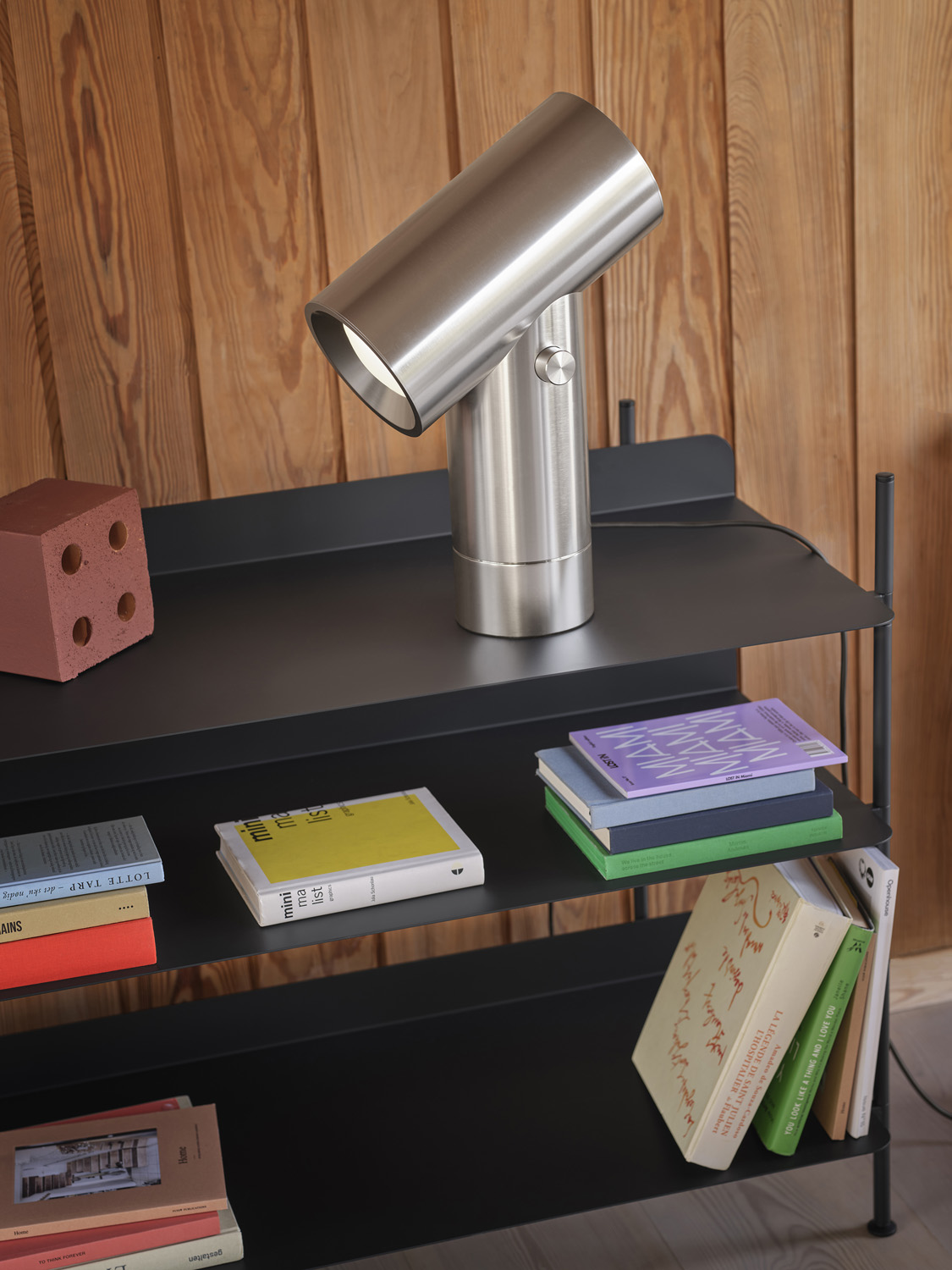 A polished aluminum lamp standing on a black compile shelving system. This 45 cm beam lamp offers a striking visual balance, perfect for enhancing contemporary workspaces or living areas.