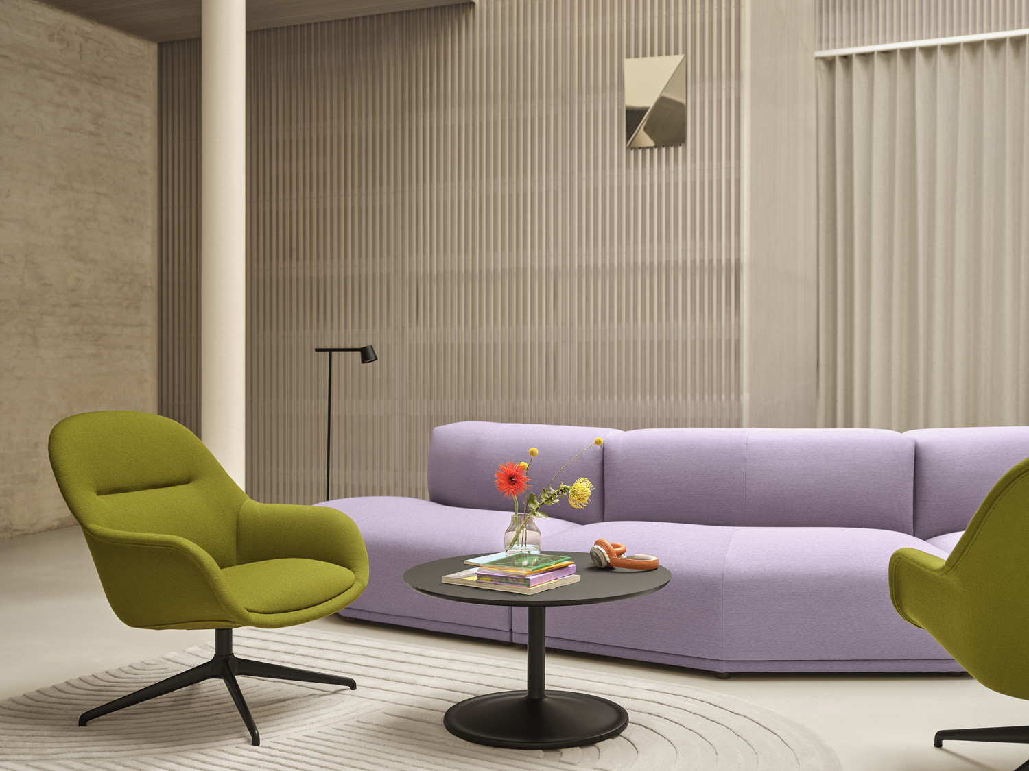 A purple sofa and two green armchairs placed in a big modern and minimalistic room together with a coffee table in black. A mirror is placed in the back of the room. 