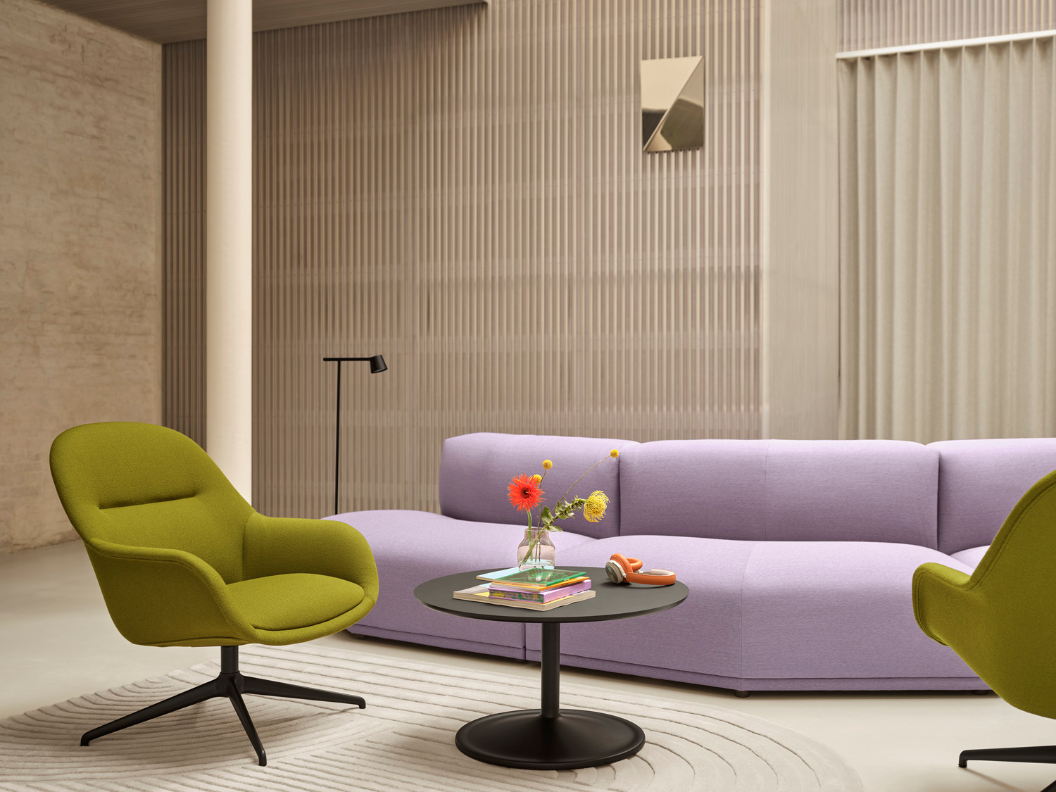 A purple sofa and two green armchairs placed in a big modern and minimalistic room together with a coffee table in black. A mirror is placed in the back of the room. 