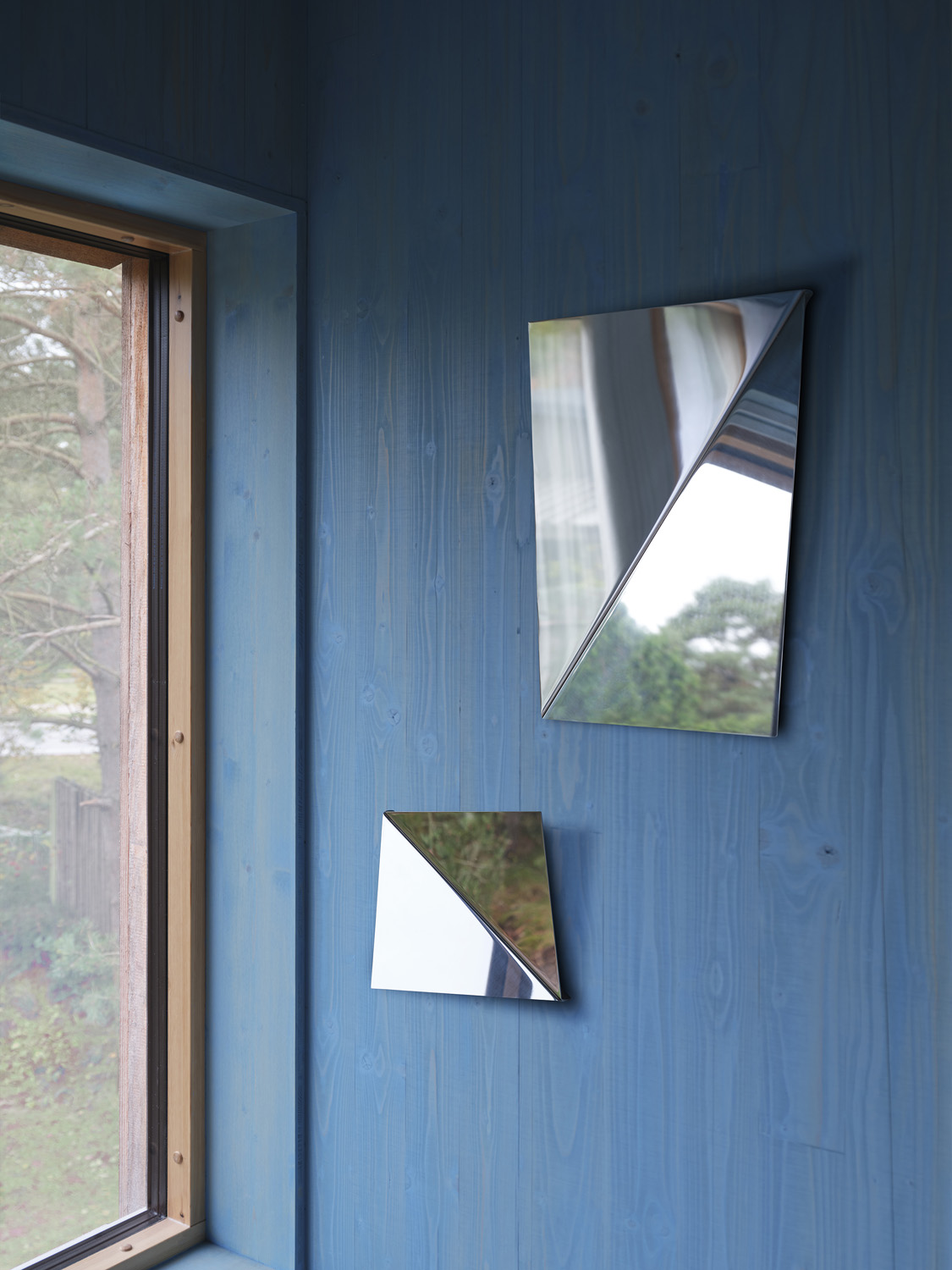 Two reflecting Gaze Mirrors hangning on a blue wall. With their gently angled surfaces they create a new perspective on a wall-mounted image mirror. Its clean expression and precise and neat detailing gives the mirror its strong character. 