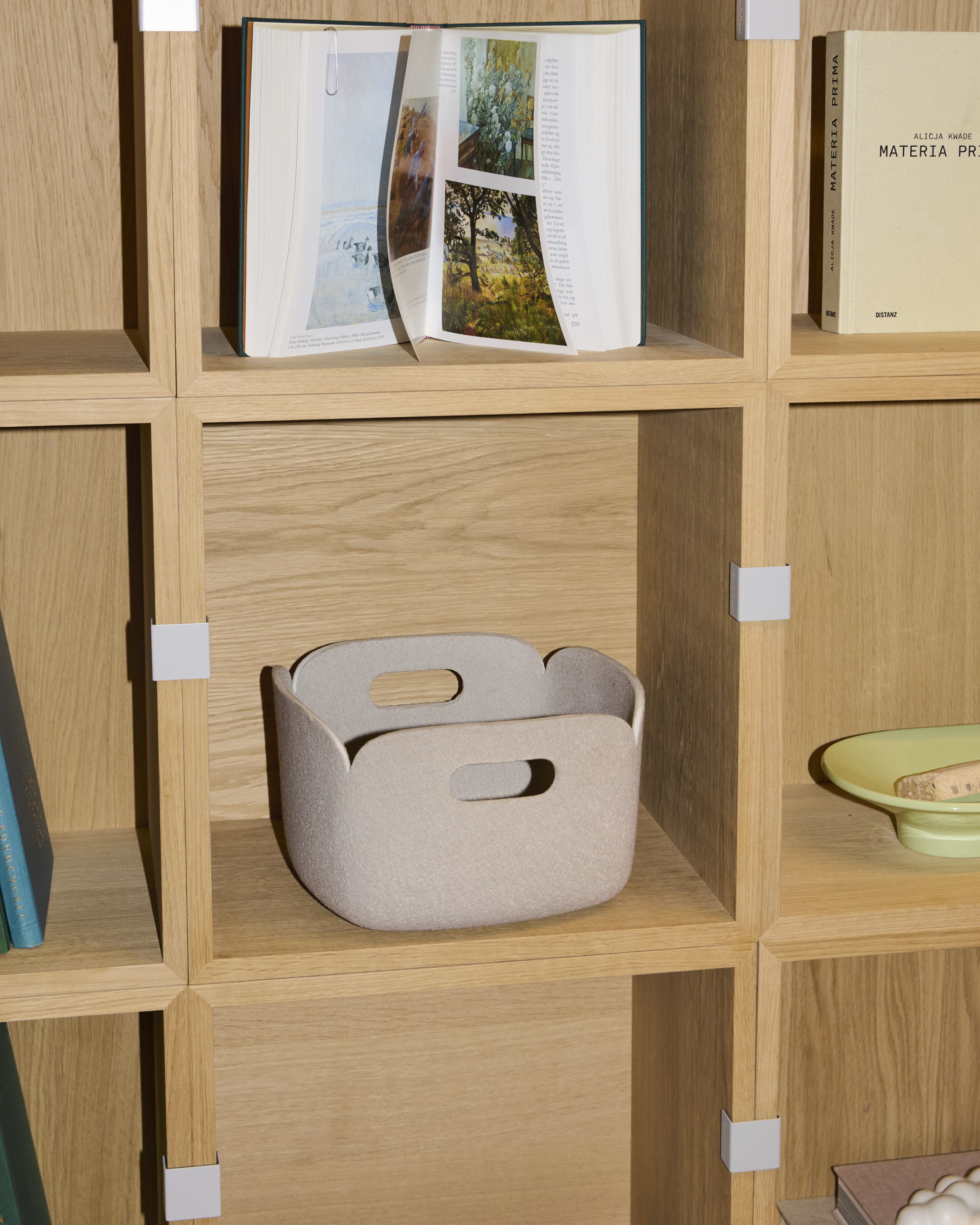 Muuto Restore Basket in Sand color. The basket is showcased against a Stacked Bookcase in oak, highlighting its soft, minimalist design and functional storage capabilities. The material appears to be recycled felt, providing a durable yet sustainable touch to the contemporary aesthetic.