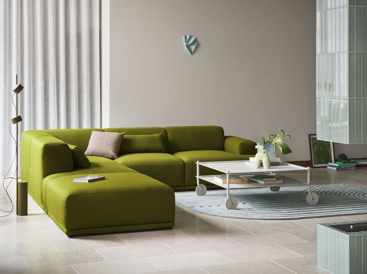 A Muuto living area featuring the Flow coffee table in grey, paired with the Connect Soft sofa with Vidar 956 upholstery. The scene includes a Revelo rug in sage green, a Post floor lamp in brown green, and a Mingle cushion. The combination of modern furniture and accents creates a stylish and inviting space.