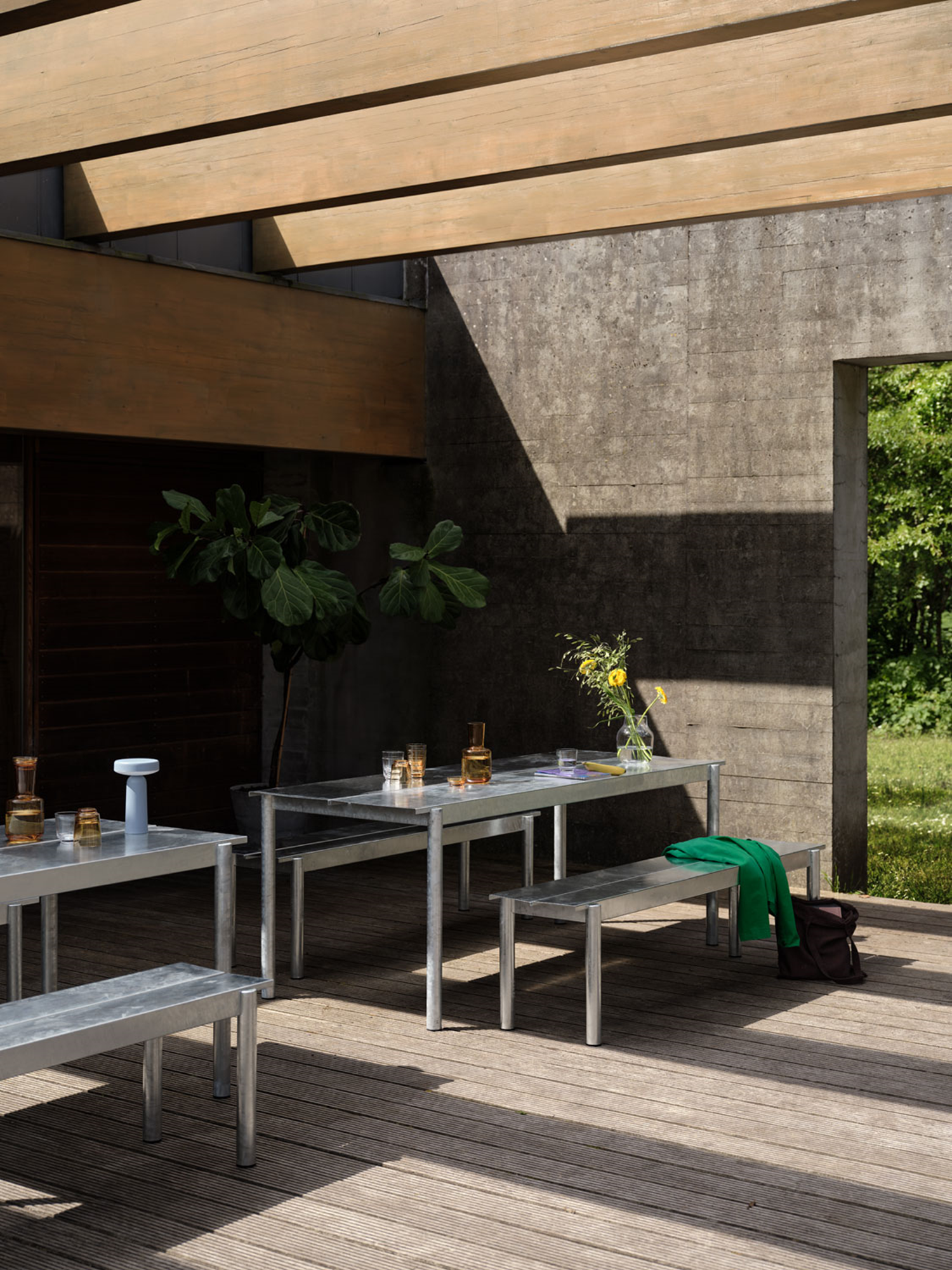 A Muuto setup featuring a Linear steel bench in warm galvanized steel and Ease portable lamp in light blue. The scene includes a Raise carafe and glasses in burnt orange, along with a Silent vase. The combination of metal furniture and elegant accessories creates a stylish and inviting arrangement.