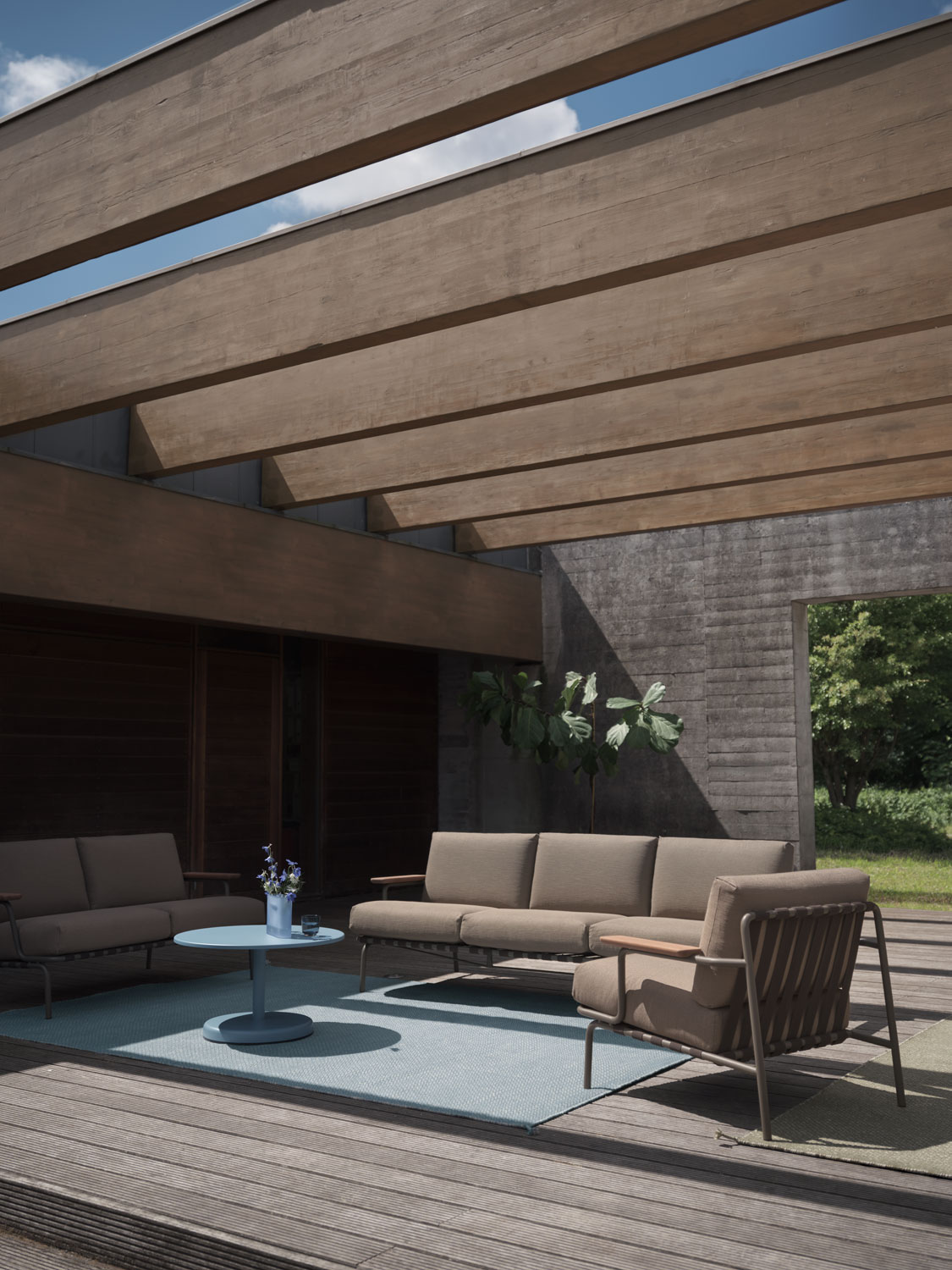 A Muuto living space featuring a Settle sofa 2 and 3 seater and lounge chair with ribbed weave 5 upholstery in taupe. The arrangement includes a light blue Ply rug made from recycled polyester and a Ridge vase 16 cm in glass. The combination of soft textiles and modern accents creates a stylish and inviting environment.