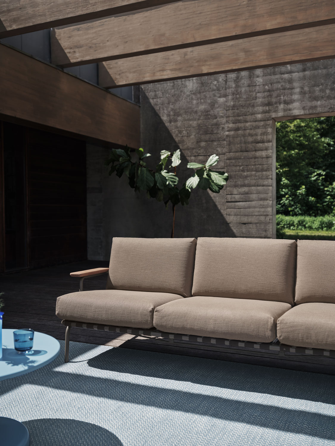 A Muuto living space featuring a Settle 3-seater sofa with ribbed weave upholstery in taupe. The sofa is complemented by a light blue Ply rug made from recycled polyester. The combination of the textured sofa and the soft rug creates a warm and modern outdoor space.
