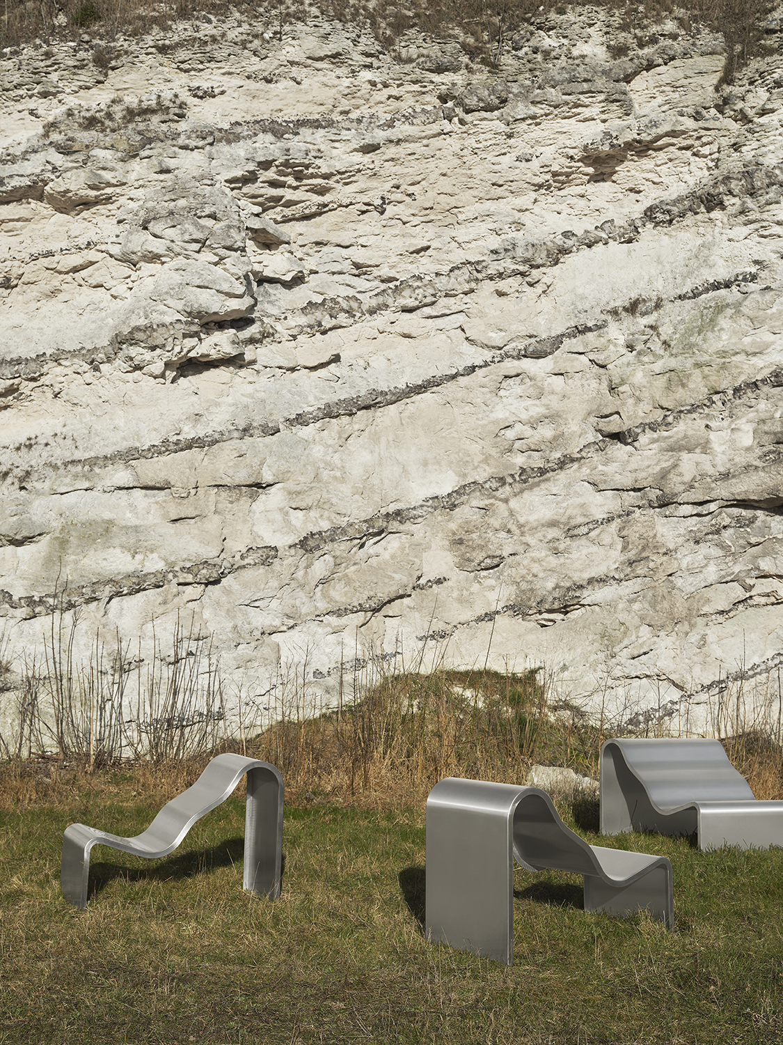 Three architectural metal chairs with a wavy, sculptural design are placed outdoors on a grassy surface, creating a striking contrast between the industrial form and natural setting.