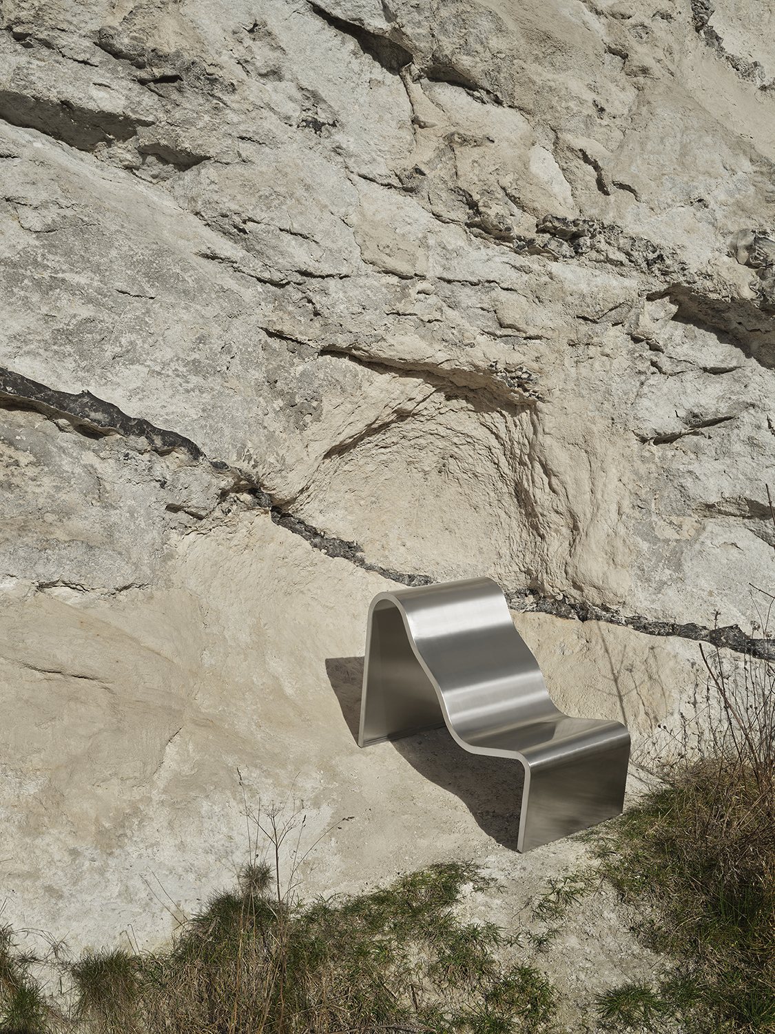 An architectural metal chair with a wavy, sculptural design is placed outdoors on a grassy surface, creating a striking contrast between the industrial form and natural setting.