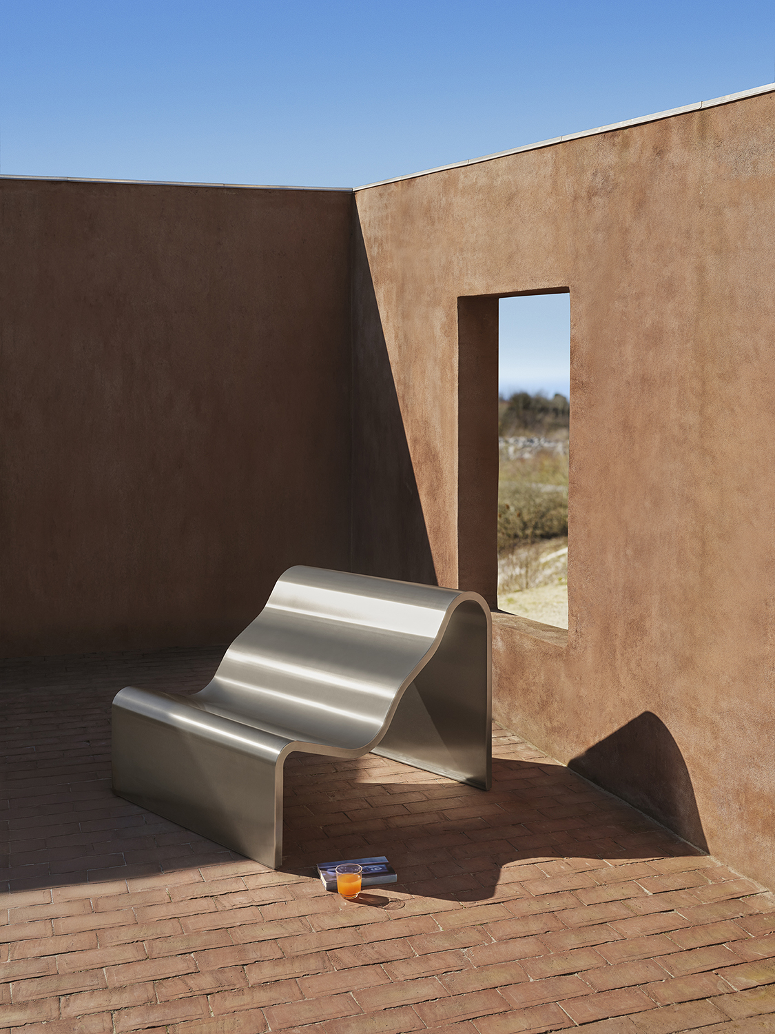A architectural metal bench with a wavy, sculptural design is placed outdoor. A glass in a clear color is placed beside the bench. 