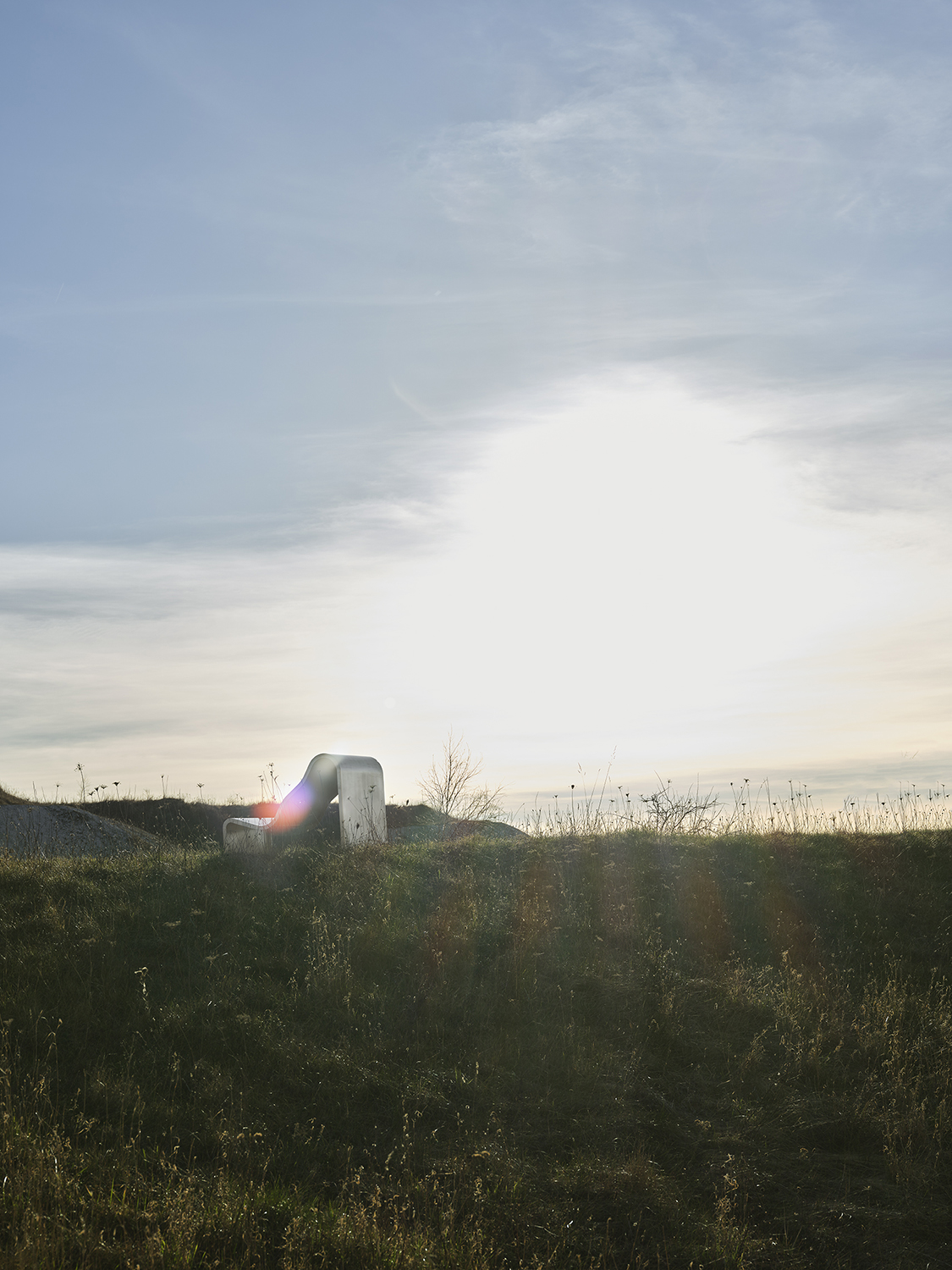 An architectural metal bench  with a wavy, sculptural design is placed outside in the sun on the grass. 