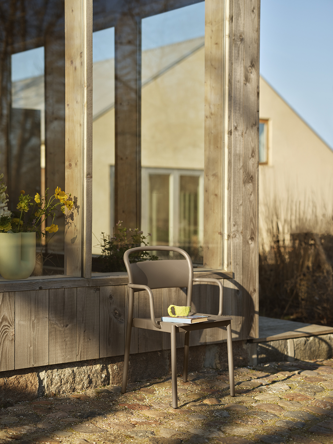 A linear steel armchairin  taupe placed outside in the evening sunlight. A Light green Kink vase in placed behind the chair creating a cozy setting. 