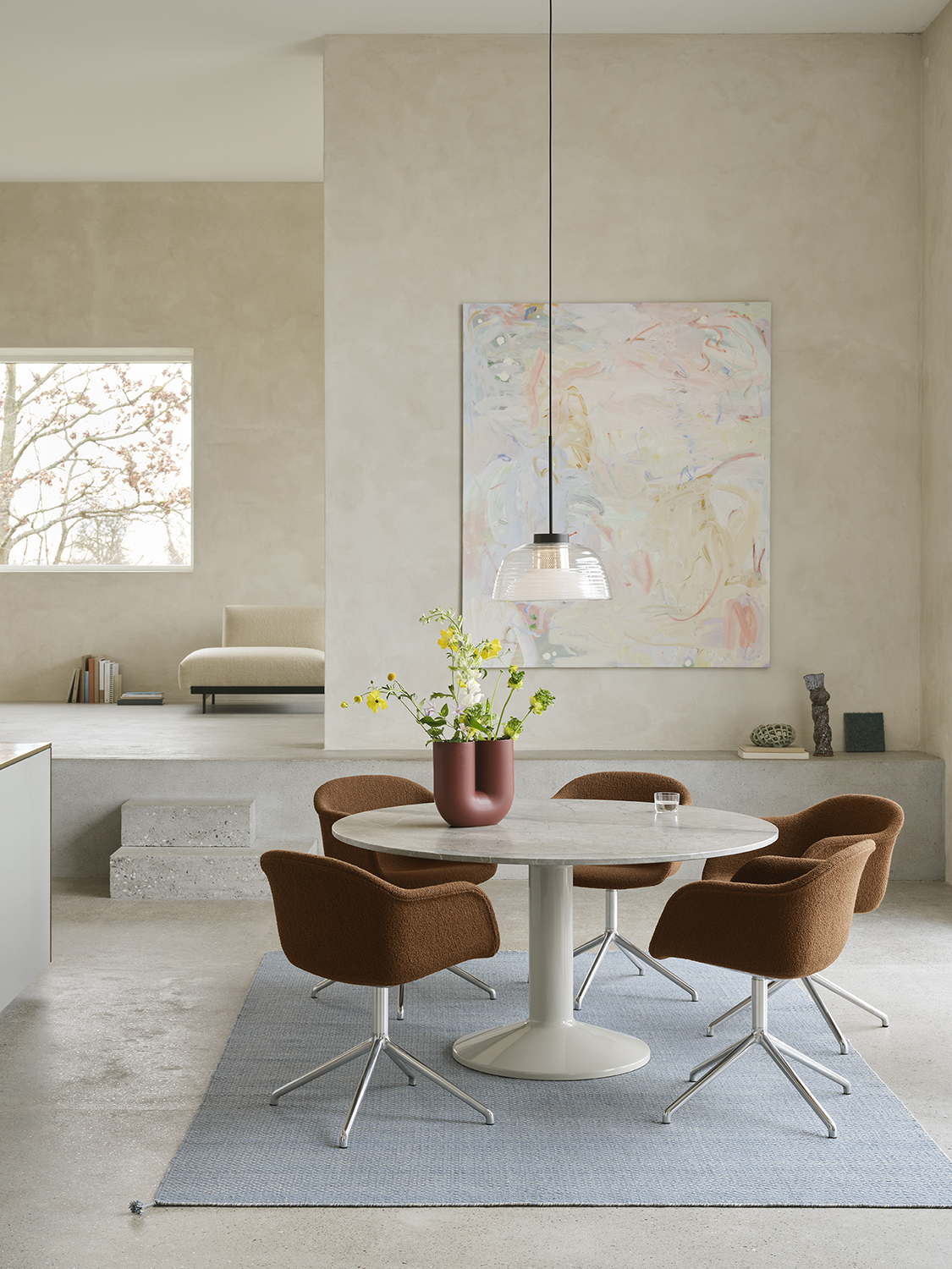 A round grey marble dining table sits in a minimalistic dining room with brushed stone walls and floors. Soft brown dining chairs are arranged around the table, with a light blue rug beneath adding a pop of color. A glass pendant lamp hangs above the table, completing the serene and modern atmosphere.