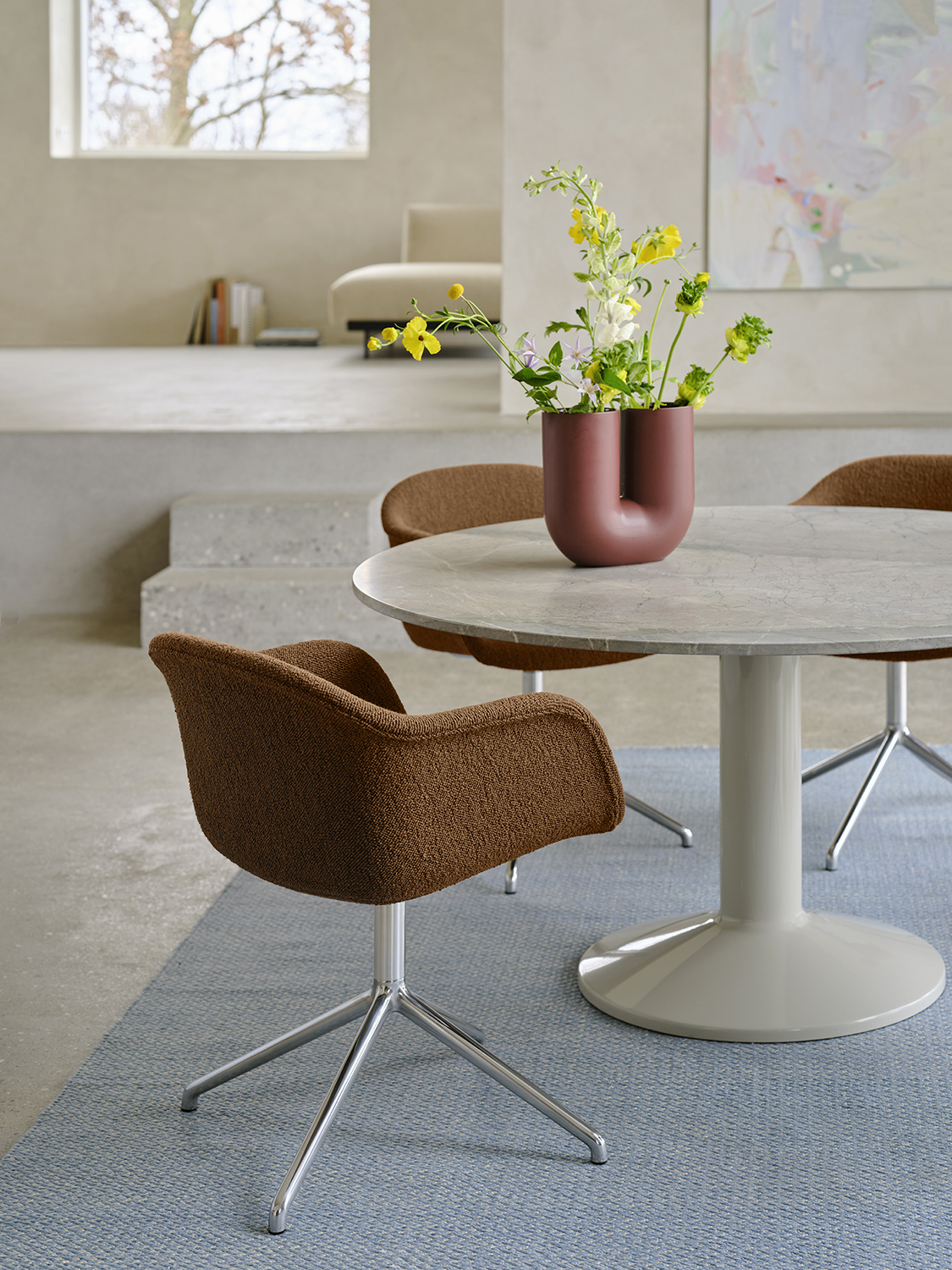 A close-up of a round grey marble dining table in a minimalistic dining room with brushed stone walls and floors. A soft brown dining chair is placed at the table, with a light blue rug underneath adding a pop of color. A sculptural vase with flowers sits on the table, enhancing the serene and modern atmosphere.