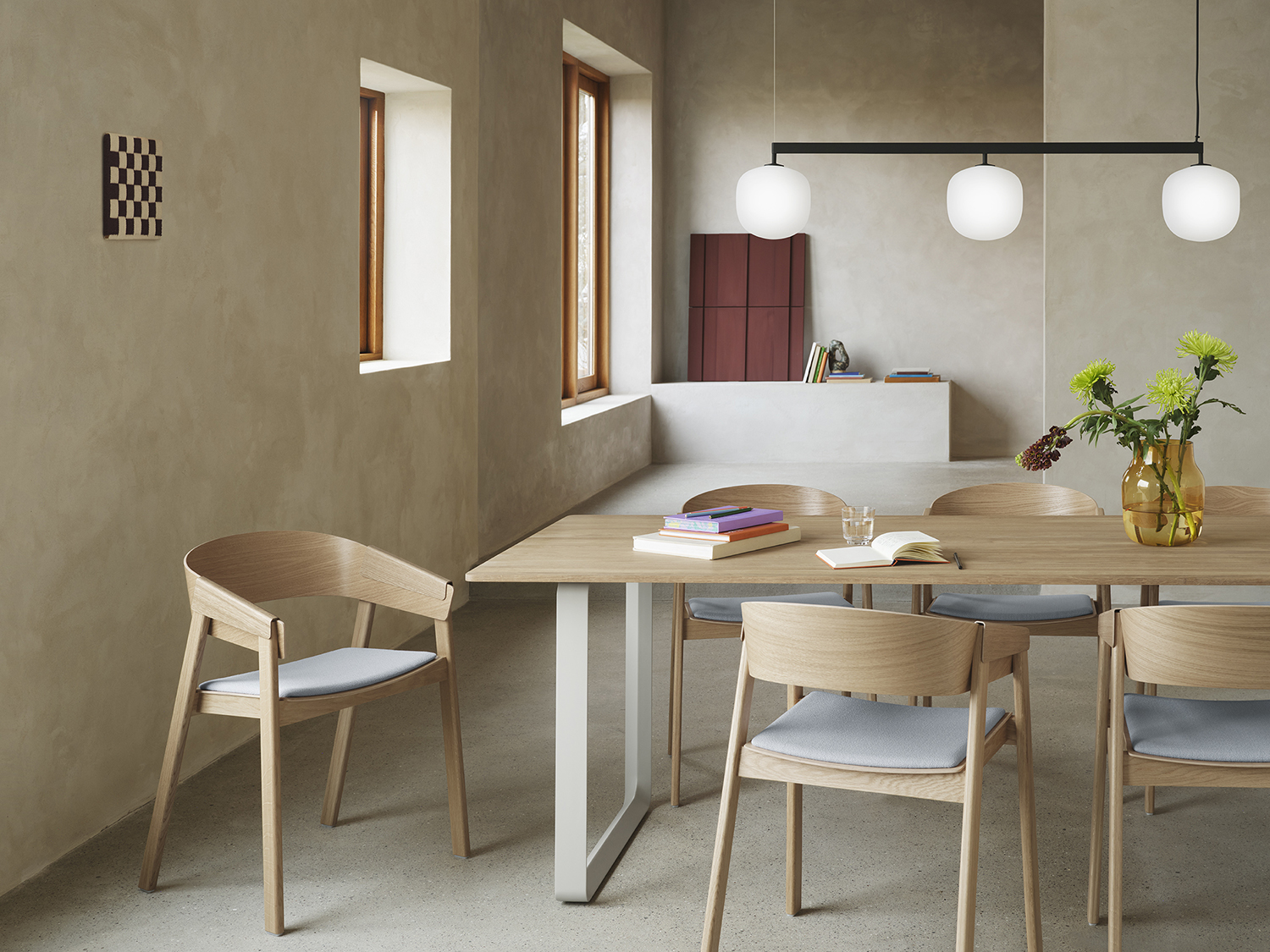 Modern wooden dining chairs are arranged around a wooden dining table in a minimalistic kitchen lounge area with brushed stone surfaces. A sleek, modern pendant lamp hangs above the table setting, enhancing the clean and natural aesthetic.