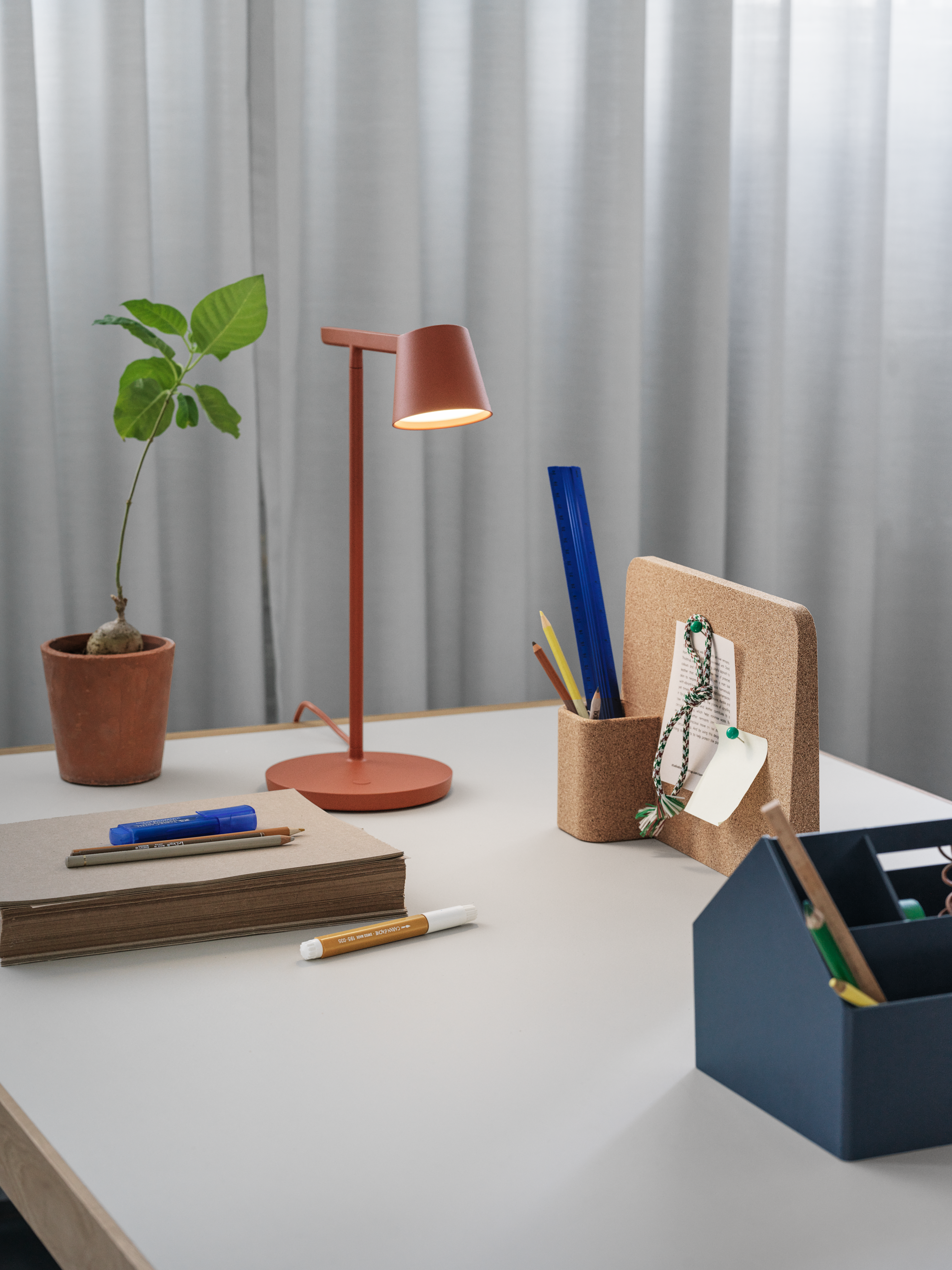 Workspace setting featuring the Workshop Table 140 cm in grey linoleum, Tip Table Lamp in copper brown, Sketch Toolbox in midnight blue, and Story Pinboard. A functional styled setup by Muuto.