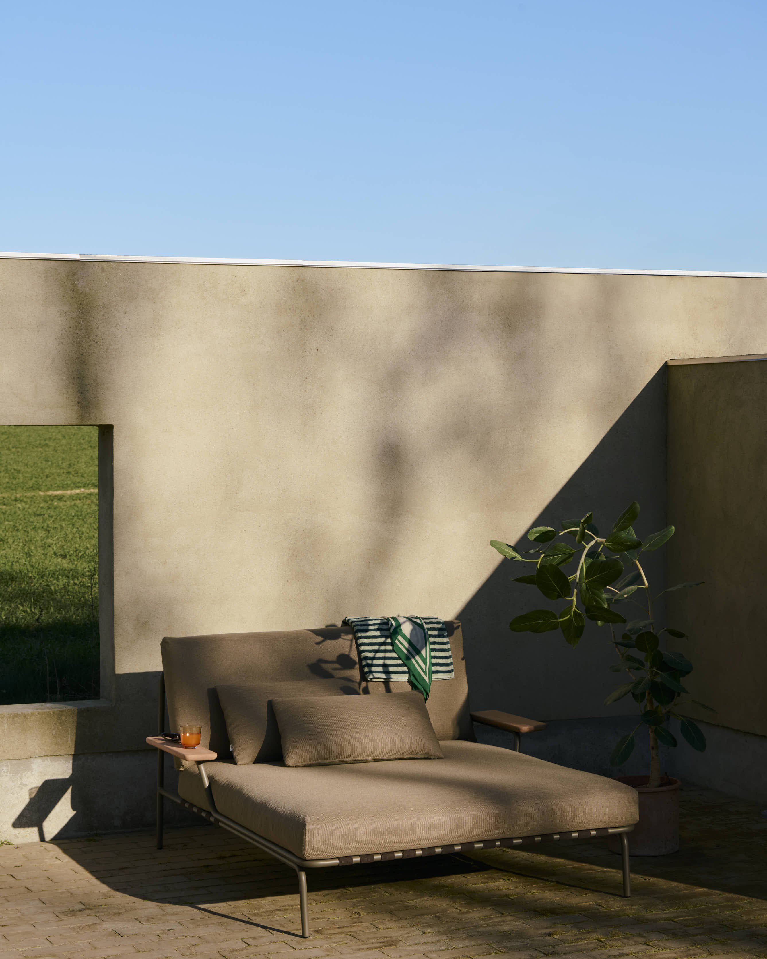 A modern, sleek lounge bed in a dark sand-colored fabric with an armrest, positioned outdoors and bathed in the warm light of the afternoon sun.