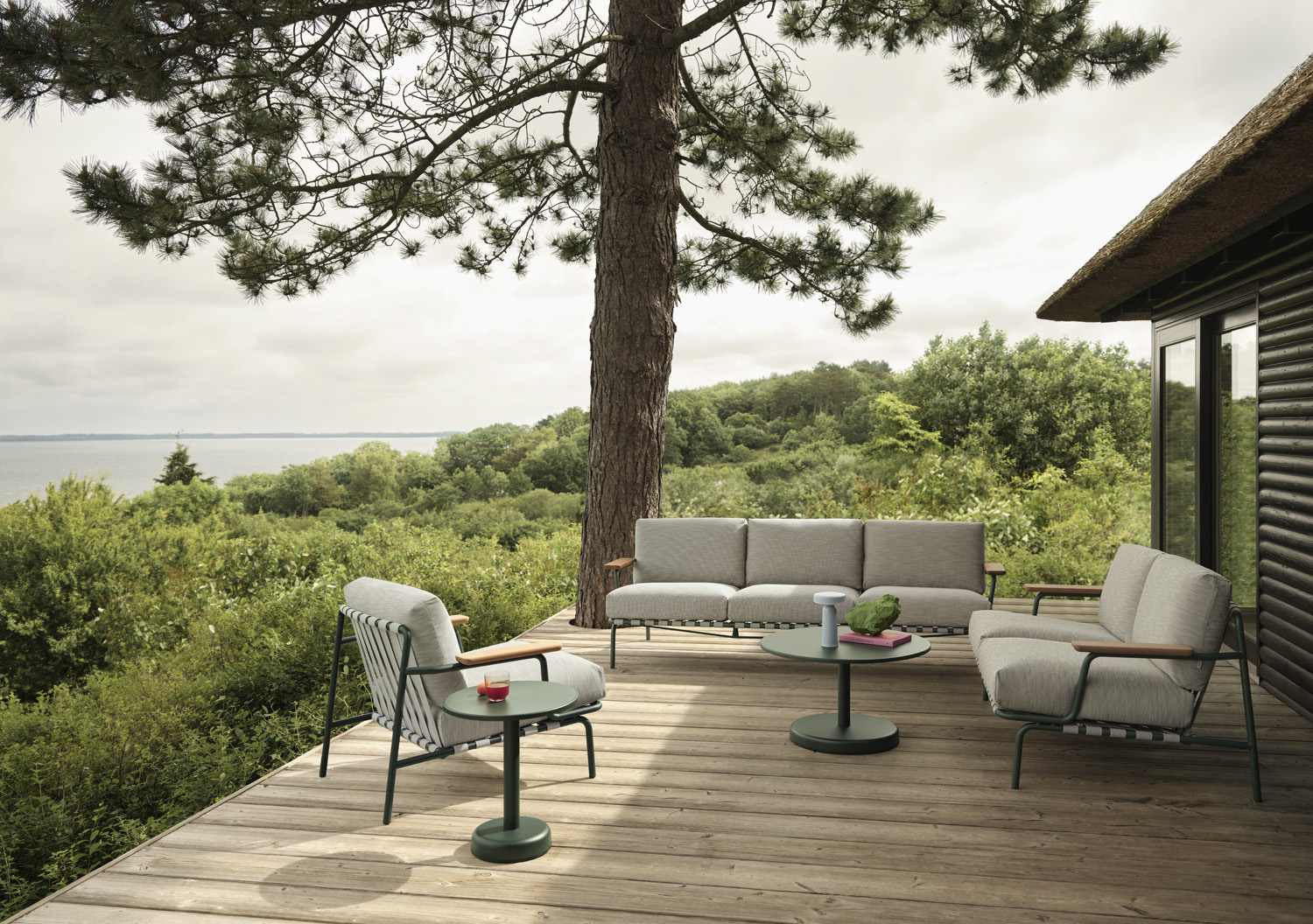Spacious outdoor deck featuring modern furniture including a light grey sofa set and a striped armchair, arranged around dark green round coffee tables with a view on the ocean. 