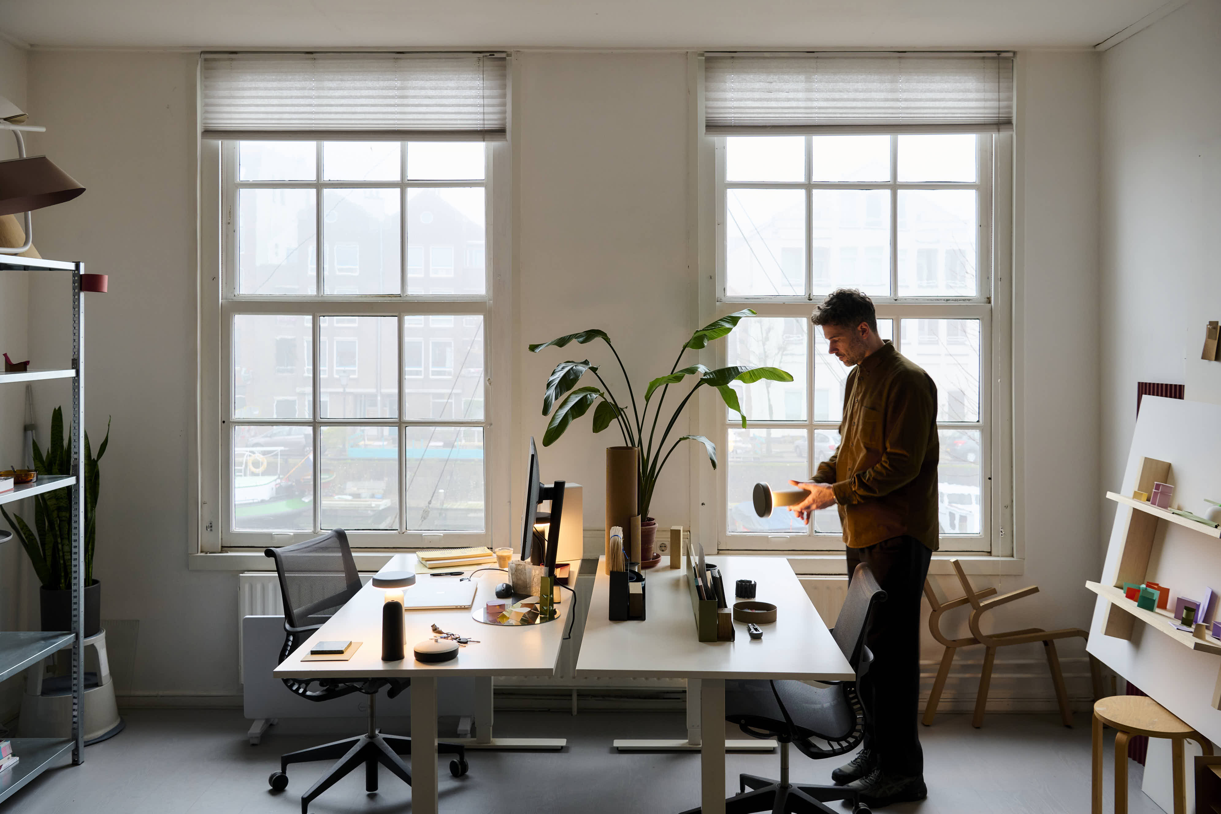 A spacious office with large windows, two desks, and Johan holding a Muuto Ease portable lamp. The workspace is decorated with plants, shelves, and design objects.
