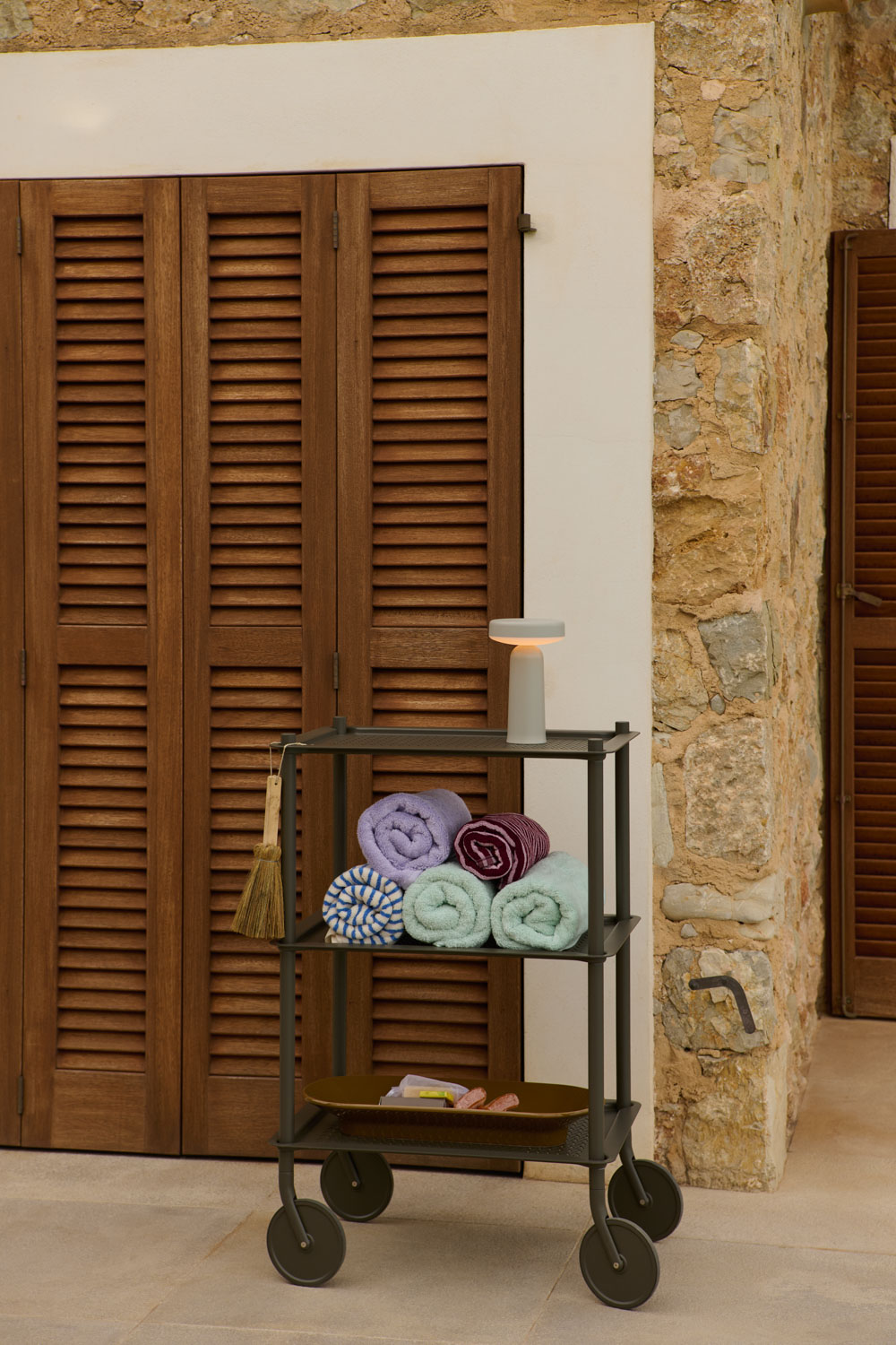A compact Ease Lamp sits on a dark green rolling cart filled with neatly rolled towels, placed outside a rustic stone house with wooden shutters.